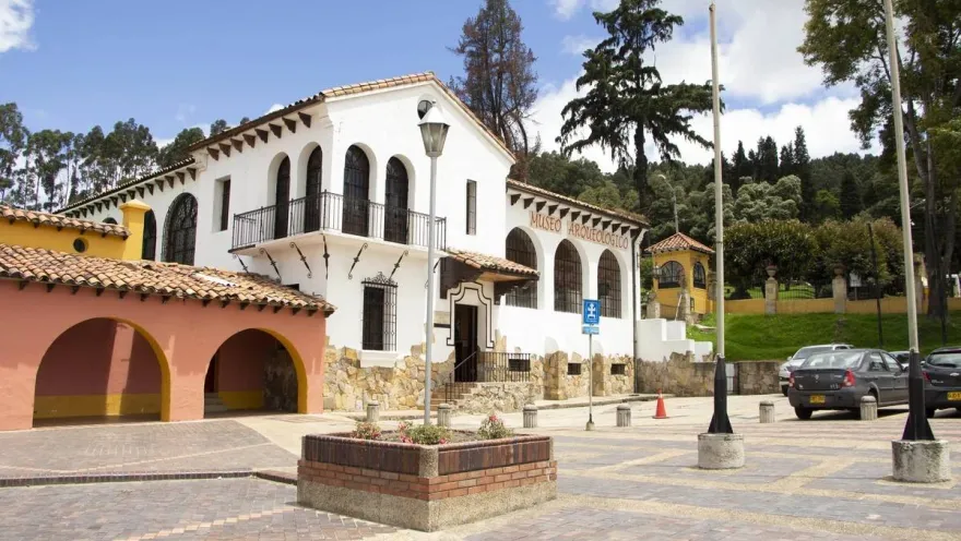 Museo Arqueológico fachada estilo colonial de coor blanco. Al frente una plazoleta con algunos vehículos estacionados y al fondo árboles altos.