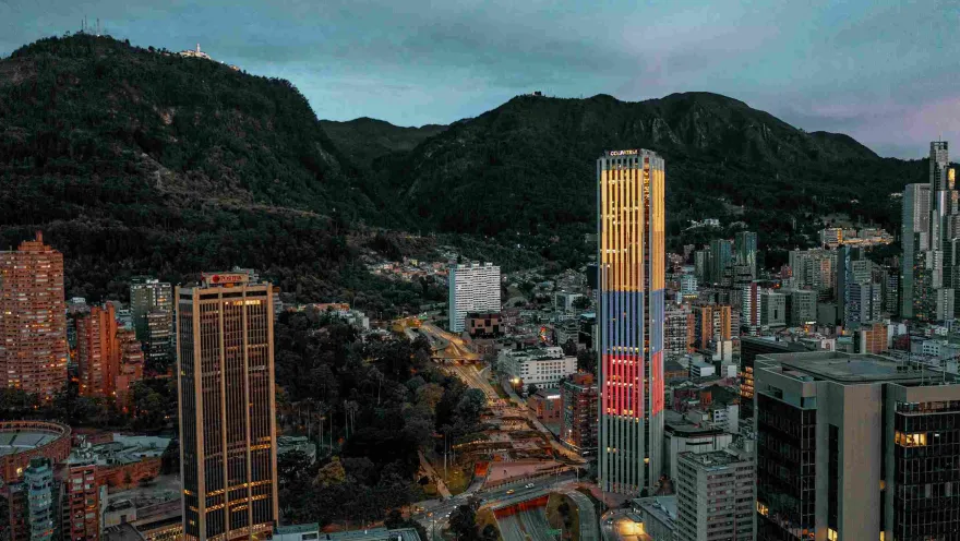 At dusk, the Colpatria Tower is illuminated with the colors of the Colombian flag, with a view from west to east showing several tall buildings and the eastern hills in the background.