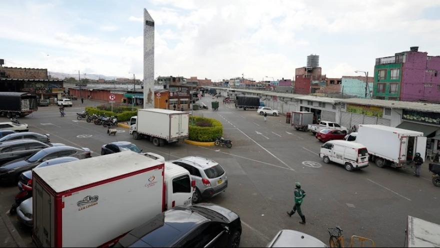 Primer plano de plaza en Corabastos se ven bodegas con locales comerciales y varios camiones alrededor. Hay un obelisco en la mitad.