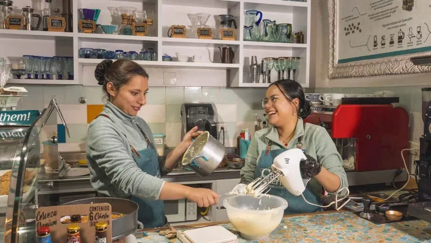 Un par de mujeres jóvenes en una cocina preparan y mezclan algo en un platón de plástico. Están sonriendo.