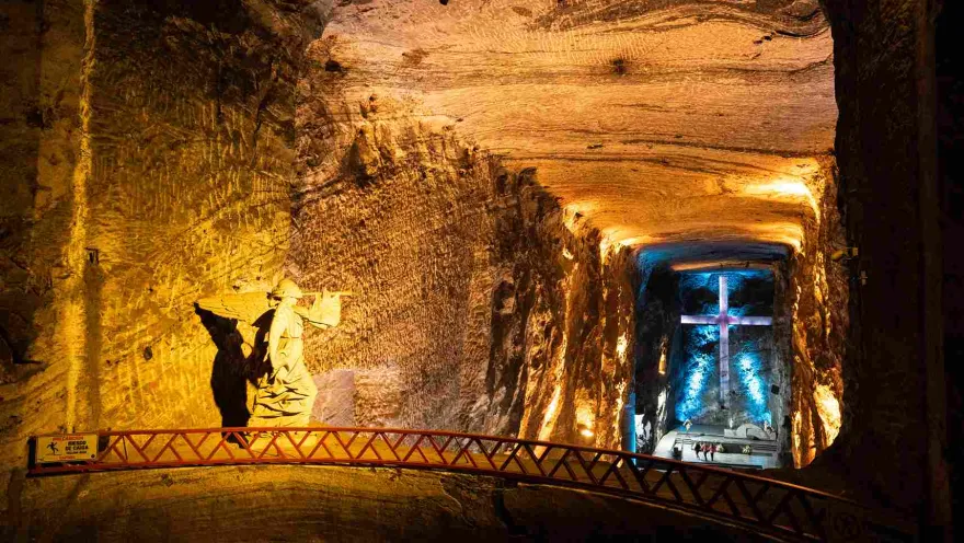 Socavón de la Catedral de Sal con una estatua de un ángel y una cruz en roca al fondo. El escenario está iluminado con luz amarilla y azul.