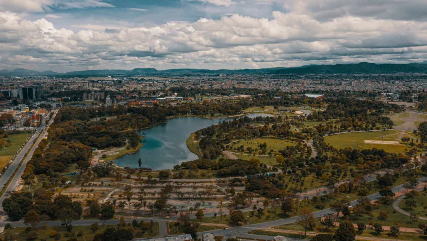 Simón Bolívar Park, a wide aerial view from a drone, looking from east to west, showing green spaces, surrounding trees, the lake in the center, and city buildings in the background. The sky is overcast.