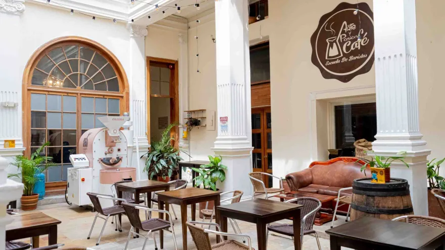 Salón principal de un café en el que se ven mesas de madera y sillas de mimbre, 3 columnas de mármol que sostienen el lugar y le dan un elegante decorado y un aviso en una pared que dice "Arte y pasión café".