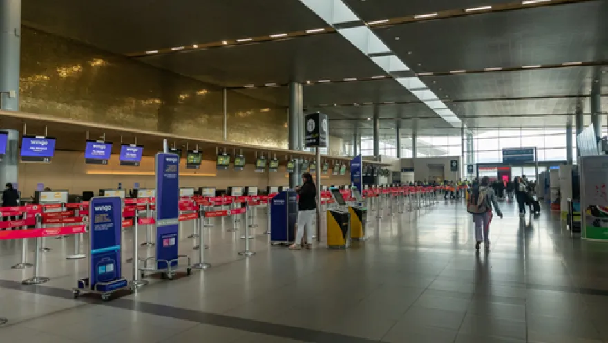 Interior of an airport showing a deep hall with several customer service areas. Some people with suitcases are walking along the front.