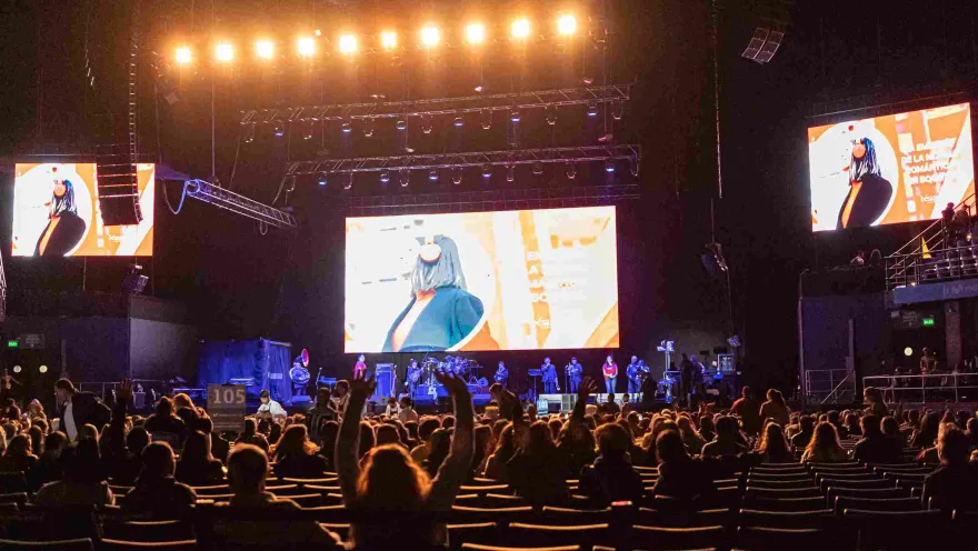 The arena in darkness with a seated audience and a performing group on stage. Three giant screens are showing a video.