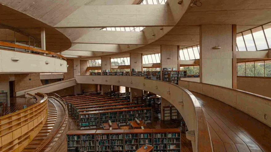 Library interior featuring circular wooden walkways, windows along the sides, and a room below with bookshelves full of books.