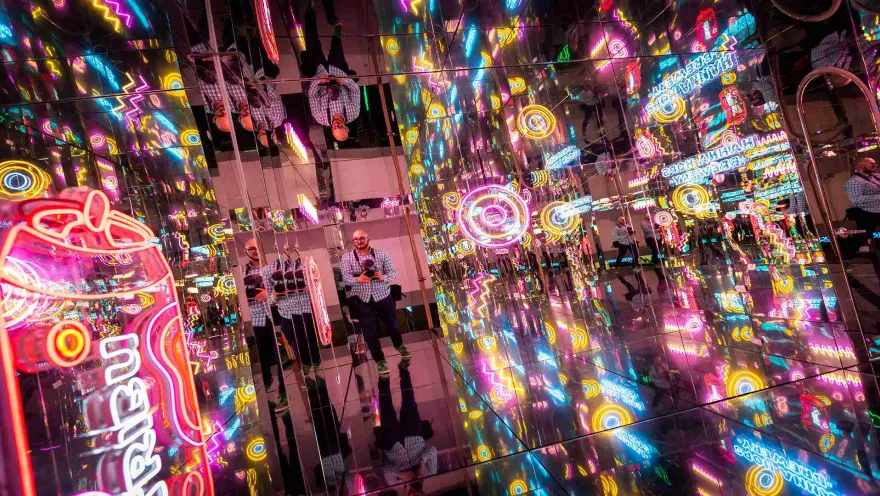 Un hombre joven y calvo con una cámara fotográfica en medio de un pasillo con paredes de espejos de cristal iluminados con luces de neón de varios colores. 