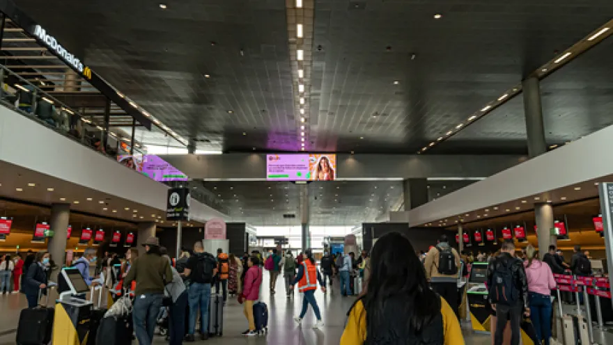 Interior of an airport showing a deep hall and some people with suitcases walking along the front.