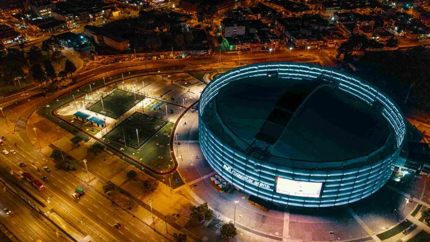 Nighttime panoramic view from above of the Movistar Arena. A couple of synthetic maids can be seen, and in the background some illuminated neighborhood buildings.