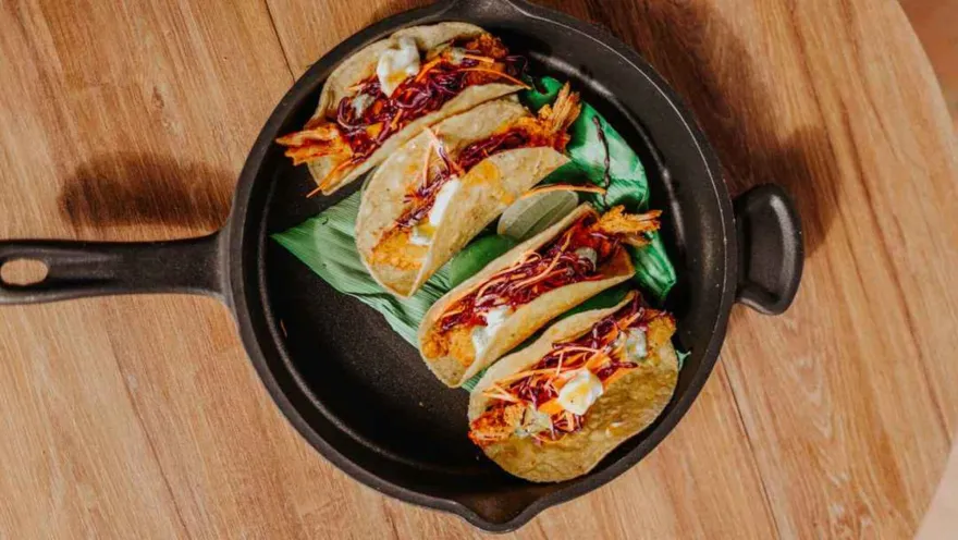 Cuatro tacos de pescado o camarones fritos con col morada, salsa cremosa y lima, servidos en una sartén de hierro fundido sobre una tabla de madera. Los tacos están envueltos en hojas verdes para presentación.