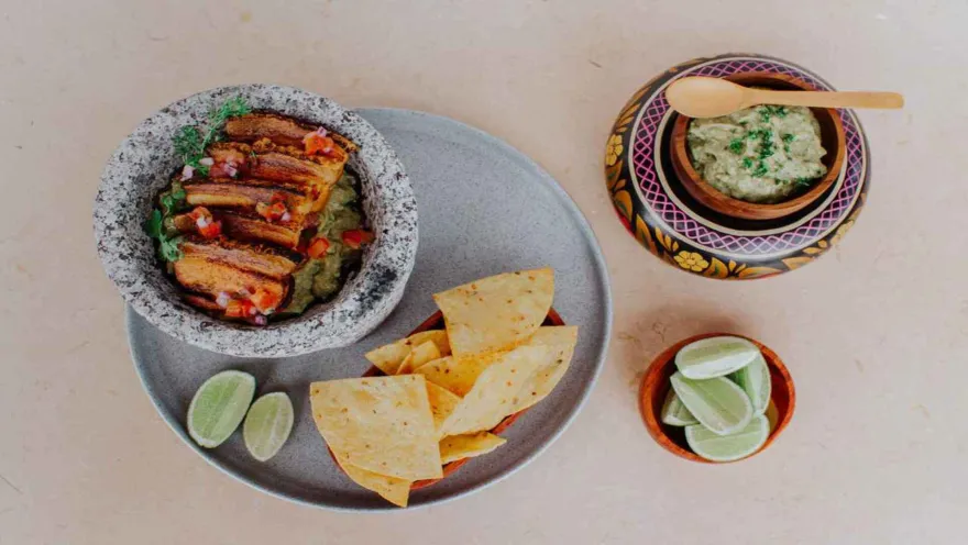 Un plato de chicharrón con guacamole en un molcajete de piedra, acompañado de totopos de maíz, rodajas de lima y un recipiente adicional de guacamole con una cuchara de madera, todo sobre una superficie clara.