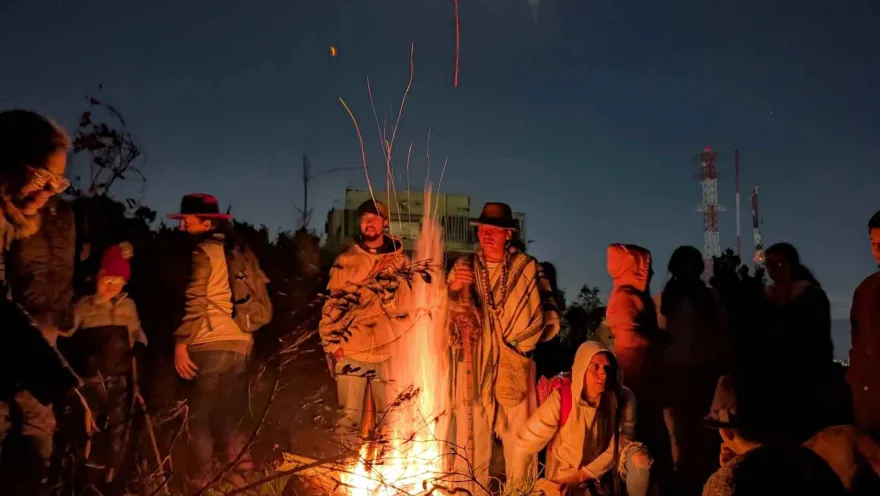 Grupo de personas entre jóvenes y adultos de noche, con chaquetas, reunidos alrededor de una chimenea. En el centro está una persona que parece un campesino o indigena con sombrero y ruana.