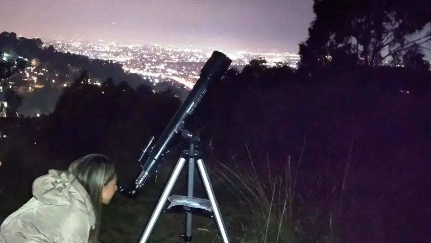 Mujer joven con una chaqueta clara observa de noche a través de un telescopio dirigido hacia las estrellas.