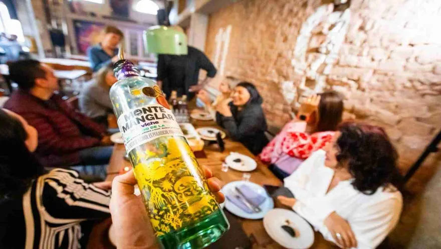 Primer plano de botella de licor en la que la etiqueta dice que es viche. Al fondo borroso se ve a un grupo de personas sentados alrededor de una mesa.