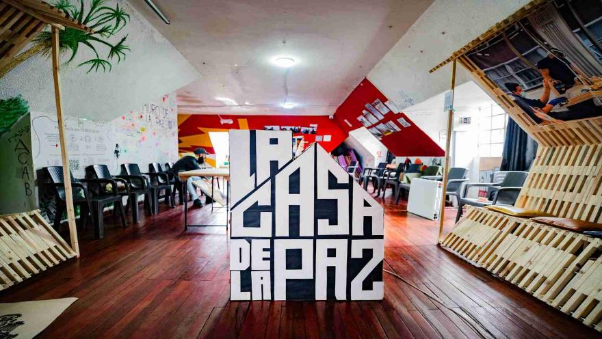 Un salón con un aviso de madera grande en el centro con la figura de una casa y el título en letras blancas y mayúsculas "LA CASA DE LA PAZ". El salón está rodeado por sillas y decoraciones de madera en las paredes.