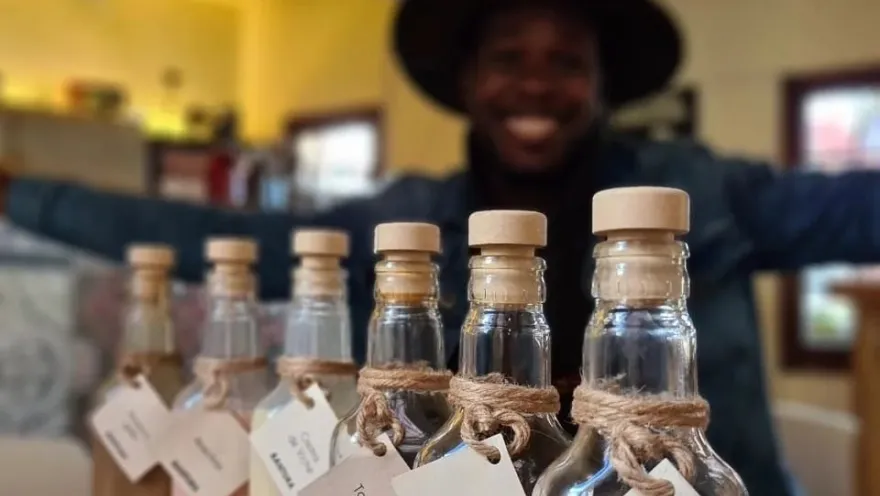  Primer plano de unas botellas de vidrio en las que resalta una leyenda que dice: "Bantura". Detrás se observa un rostro borroso de un hombre afrodescendiente con sombrero y que ríe.