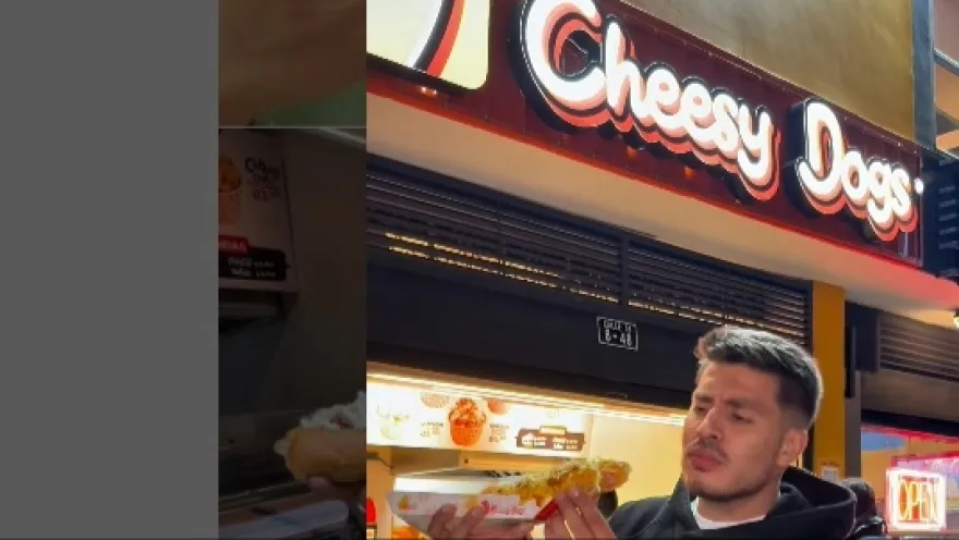 Primer plano de un hombre joven comiendo un trozo de perro caliente. Al fondo se observa un aviso con una leyenda que dice: "Cheesy Dogs".