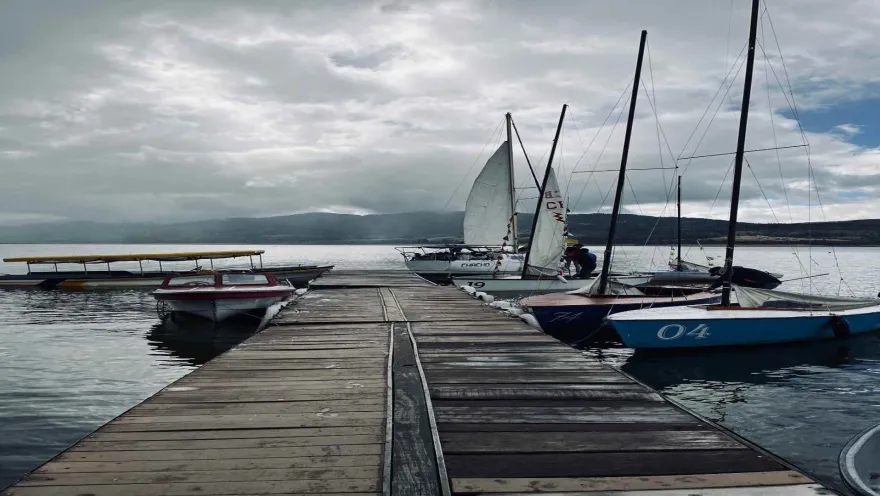 Muelle en un lago con varios veleros alrededor. El cielo está nublado.