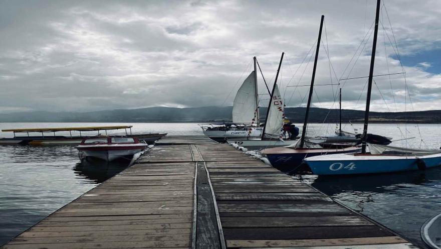 Muelle en un lago con varios veleros alrededor. El cielo está nublado.