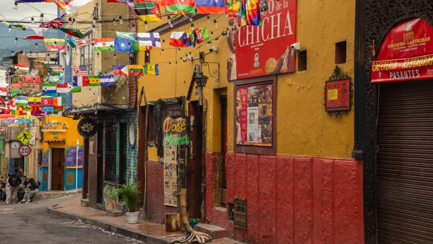 Calle con varias casas antiguas, cuyas fachadas llevan vistosos colores.Destaca un aviso con una leyenda que dice: "Chicha".