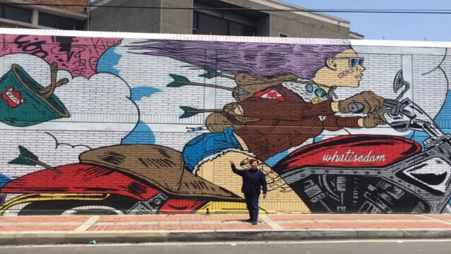  Hombre joven de pie al frente de un grafiti en el que se ve a una mujer joven que va en una motocicleta.