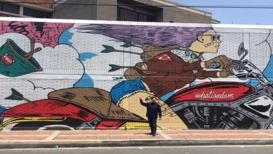  Hombre joven de pie al frente de un grafiti en el que se ve a una mujer joven que va en una motocicleta.
