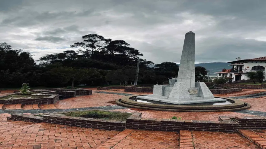 Plaza enladrillada con un obelisco no muy alto  de piedra color blanco. Al fondo se ven varios árboles frondosos.