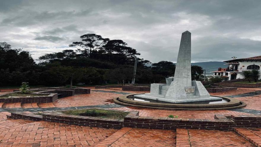 Plaza enladrillada con un obelisco no muy alto  de piedra color blanco. Al fondo se ven varios árboles frondosos.
