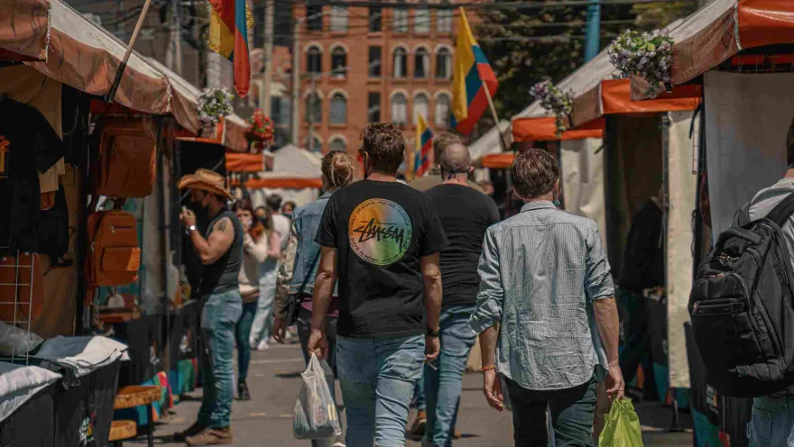 Hombres jóvenes, parecen turistas, que caminan al frente de unos toldos de comercio callejero. En los lugares se observan varias banderas de Colombia.