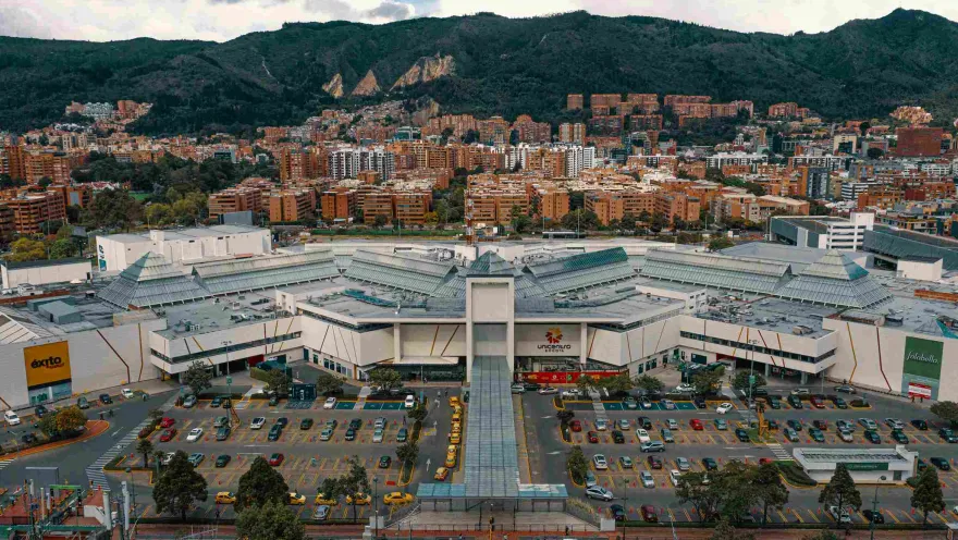  Vista desde arriba de la edificación del Centro Comercial Unicentro con su moderna estructura en concreto de color blanco, una amplia zona de parqueaderos. Al fondo se ven edificios que llegan hasta en los cerros orientales.