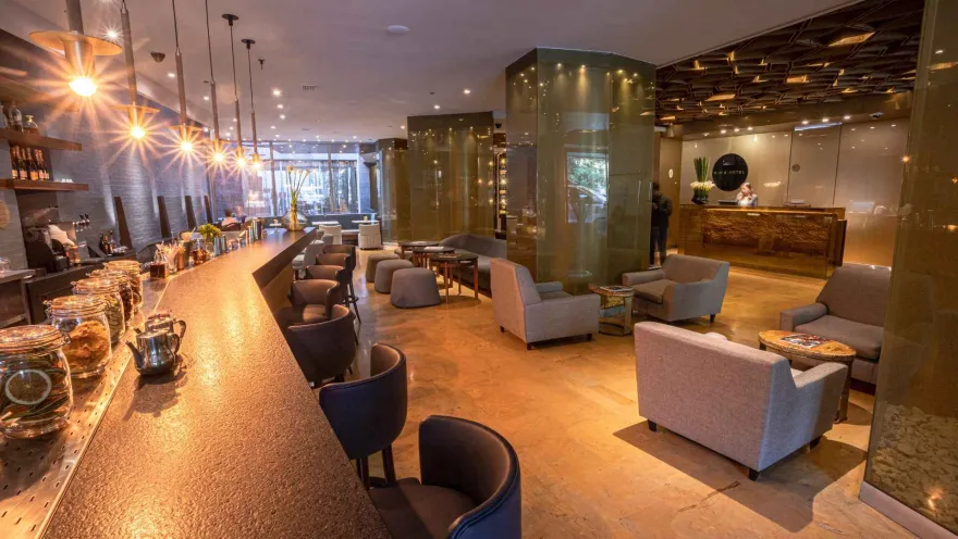 Un moderno bar de hotel con una barra iluminada, taburetes elegantes y varias áreas de asientos con sofás y sillones, creando un ambiente sofisticado y relajado.