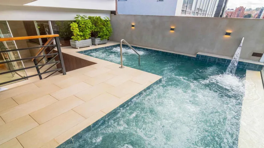 Un moderno jacuzzi exterior con chorros de agua, rodeado de baldosas claras y plantas. Las luces en la pared y una barandilla metálica completan el diseño funcional.