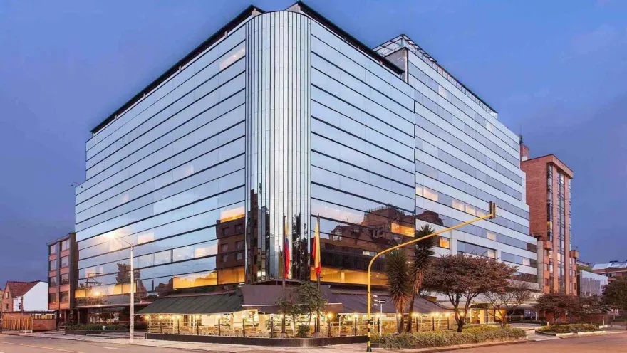  Un moderno edificio de hotel con fachada de cristal, que refleja el entorno urbano al atardecer o al anochecer. La iluminación del restaurante en la planta baja añade calidez.
