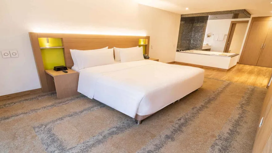 Una espaciosa habitación de hotel con una cama king-size, una cabecera de madera y un área de baño integrada con una bañera. La decoración es contemporánea.
