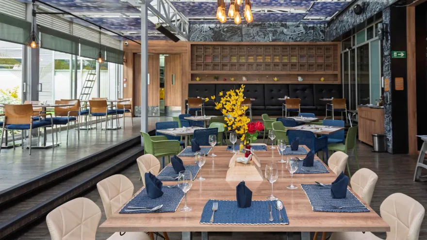 Un restaurante elegante y moderno con mesas dispuestas para cenar, sillas de colores, y una decoración única con un mural y estanterías de madera.