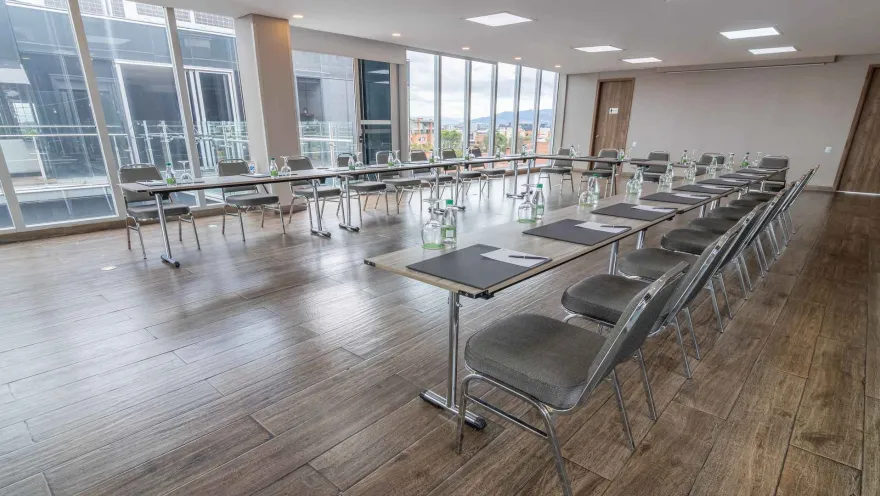  Una sala de reuniones moderna con mesas dispuestas en un gran rectángulo, sillas grises y grandes ventanales que ofrecen vistas urbanas.