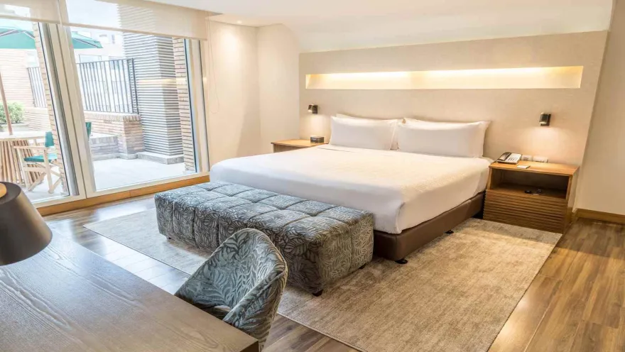  Una habitación de hotel luminosa con una cama grande, un banco acolchado, escritorio y acceso a una pequeña terraza exterior.