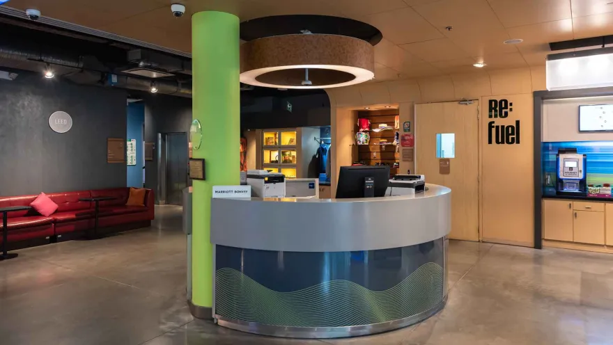 El área de recepción de un hotel Aloft con un mostrador curvo, una columna verde y acceso a la estación "Re:fuel" y un lounge.