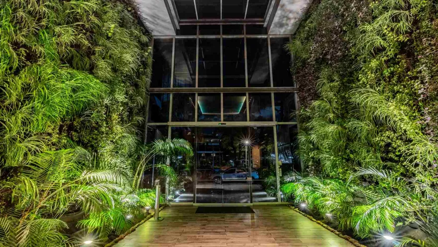 Una entrada espectacular con paredes de vegetación vertical a ambos lados y un pasillo de madera que conduce a puertas de cristal. Iluminación estratégica realza el verdor.