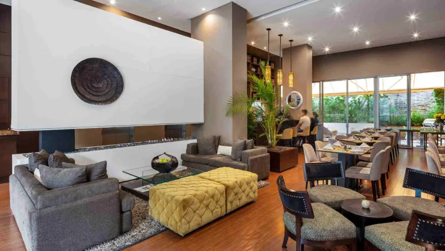 Un moderno lobby lounge que combina un área de sofás frente a una chimenea y un comedor. La decoración es sofisticada, con toques de verde y madera.