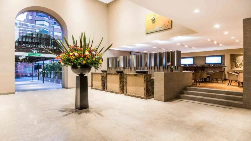 El amplio y lujoso lobby de un hotel, con una gran composición floral, mostradores de recepción de mármol y un área de bar al fondo.