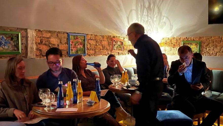 Grupo de hombres y mujeres adultos en un establecimiento, sentados alrededor de mesas con copas y bebiendo licor. Un hombre, adulto mayor les habla. 