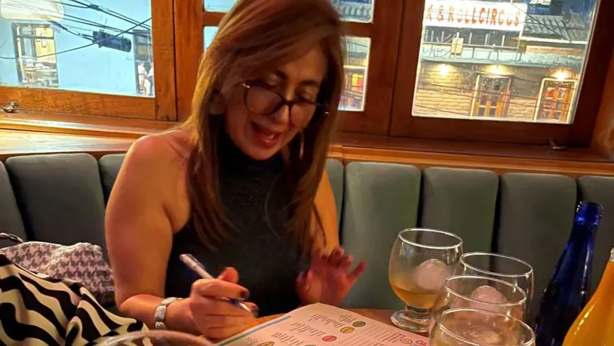  Una mujer de mediana edad con anteojos está sentada alrededor de una mesa en la que hay 4 copas y dos botellas, parece que con licor. 