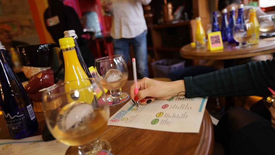Una mano de mujer escribe algo en un papel con figuras de colores. Hay varias botellas de colores y copas con un líquido que parece ser licor.