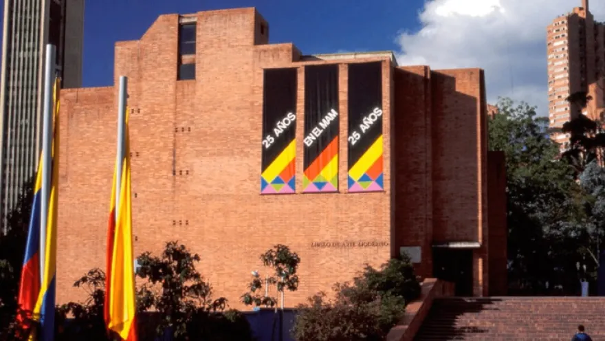 Facade of a large brick building with prominent signs in blue letters that read: “25 YEARS AT THE MAM”. A Colombian flag and a Bogotá flag are displayed to one side.