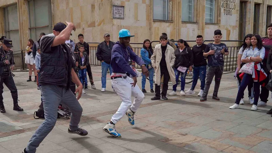 Two young men dance modern steps on a street; people watch them.