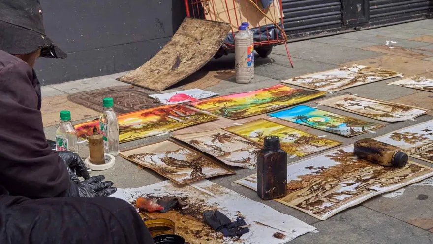 A series of paintings, seemingly freshly made, are displayed on the sidewalk. The artist can be seen from one side.