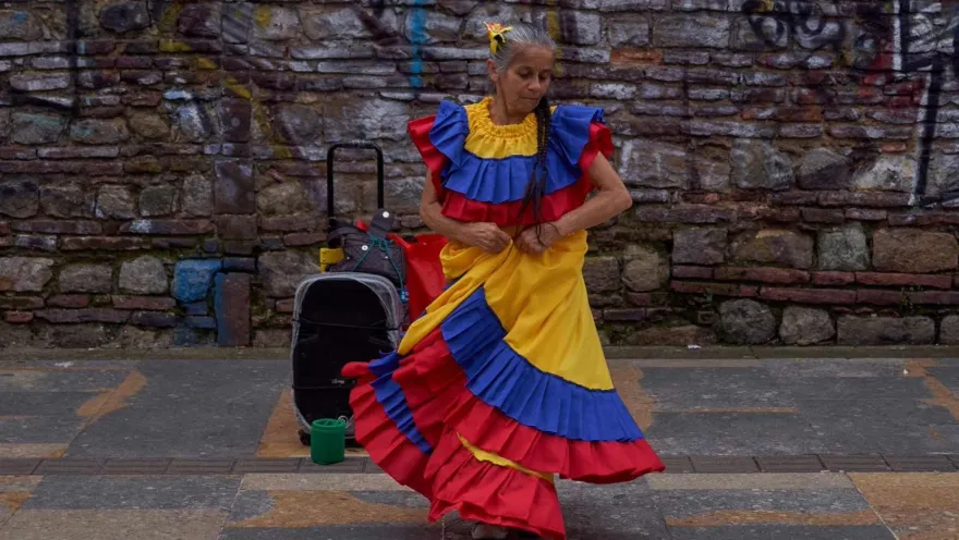 An adult woman wearing a dress in the colors of the Colombian flag dances on a street. A speaker appears to be playing the music she is dancing to.