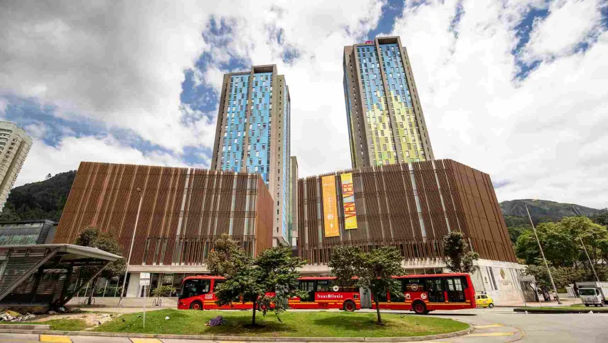 The exterior facade of a tall building constructed of concrete and wood. Behind it are a couple of high-rise residential towers, and a red TransMilenio bus is passing by. Several trees are located nearby.