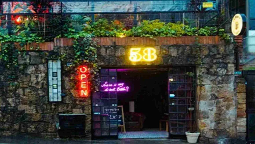  Entrada de un bar/restaurante con paredes de piedra, plantas y letreros de neón "OPEN" y "58". 